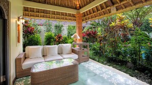 Un área de estar al aire libre con techo, que incluye un sofá de mimbre y una mesa, rodeada de exuberantes plantas tropicales y una pared decorativa.
