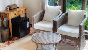 Una silla de mimbre con un cojín blanco, una mesita redonda de mimbre y una mesita auxiliar de madera con cajones y una cesta.