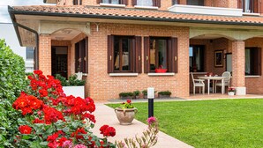 Una casa de ladrillo de dos pisos con un porche cubierto, césped y flores rojas al frente.