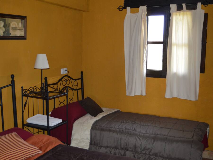 Una habitación con cama, una mesita de noche con lámpara, una ventana con cortinas y un cuadro en la pared.