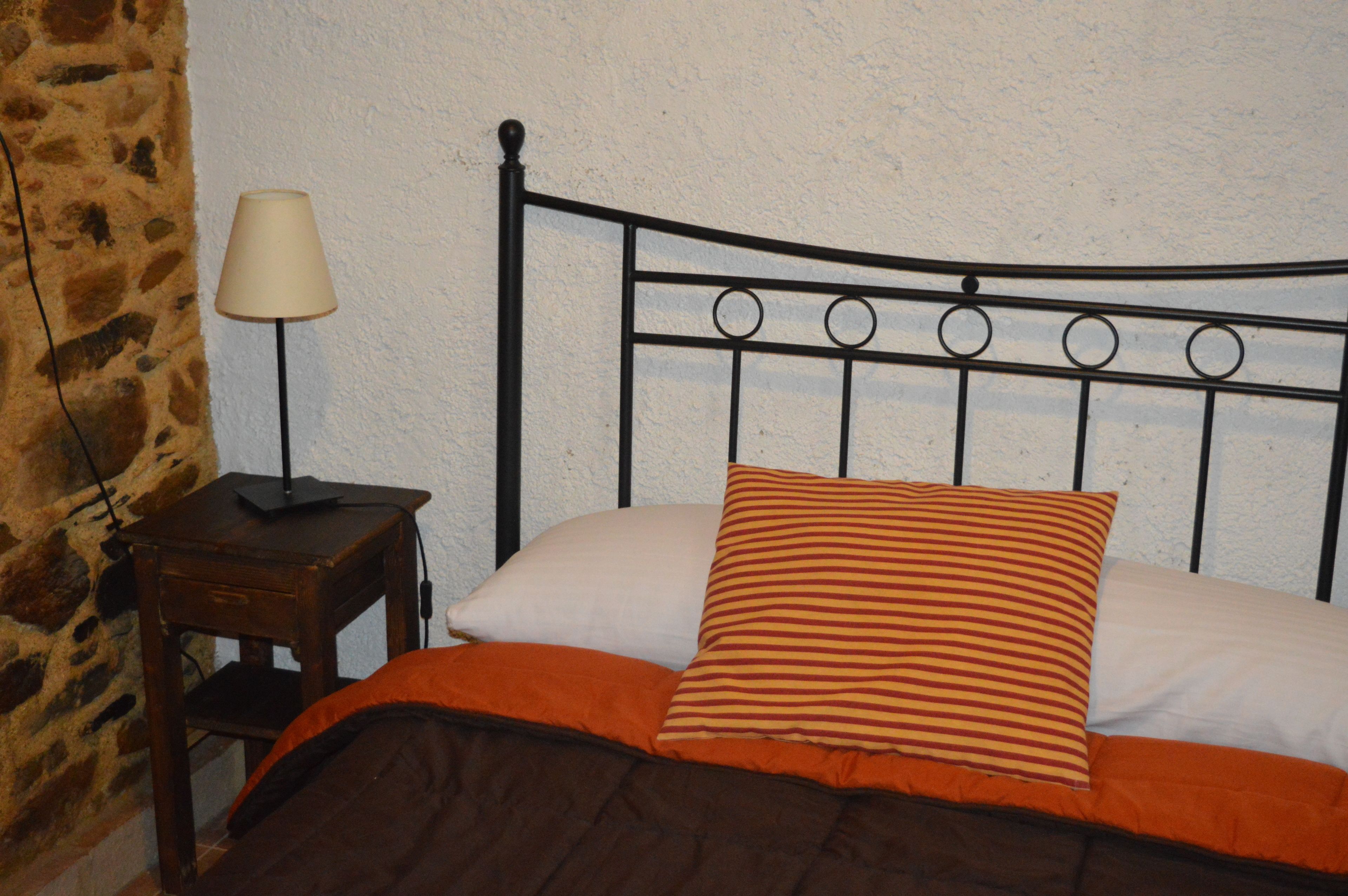 Una cama con cabecera de metal, una lámpara sobre una mesita de noche de madera y una pared de piedra.