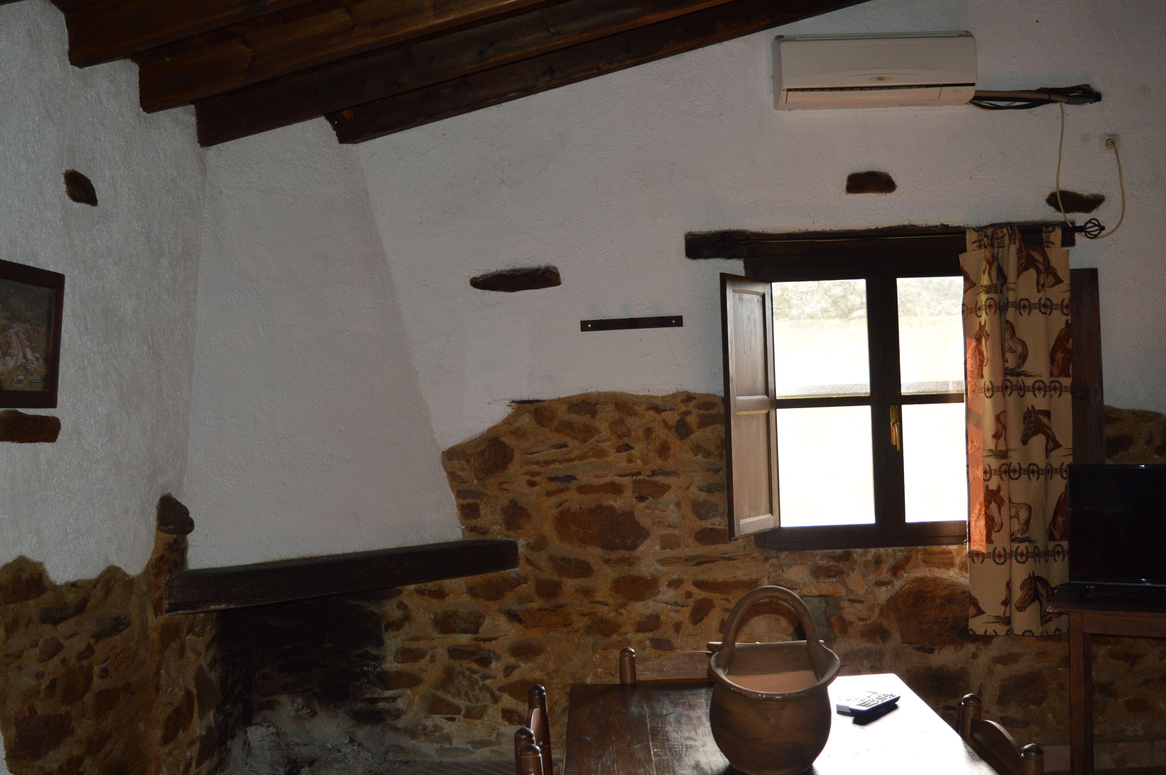 A rustic room with a stone wall, a wooden table with a kettle, a chair, a window with curtains, and a television.