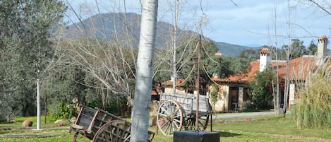 A rustic outdoor setting with a wooden cart, a stone building with a red-tiled roof, and a backdrop of trees and mountains.