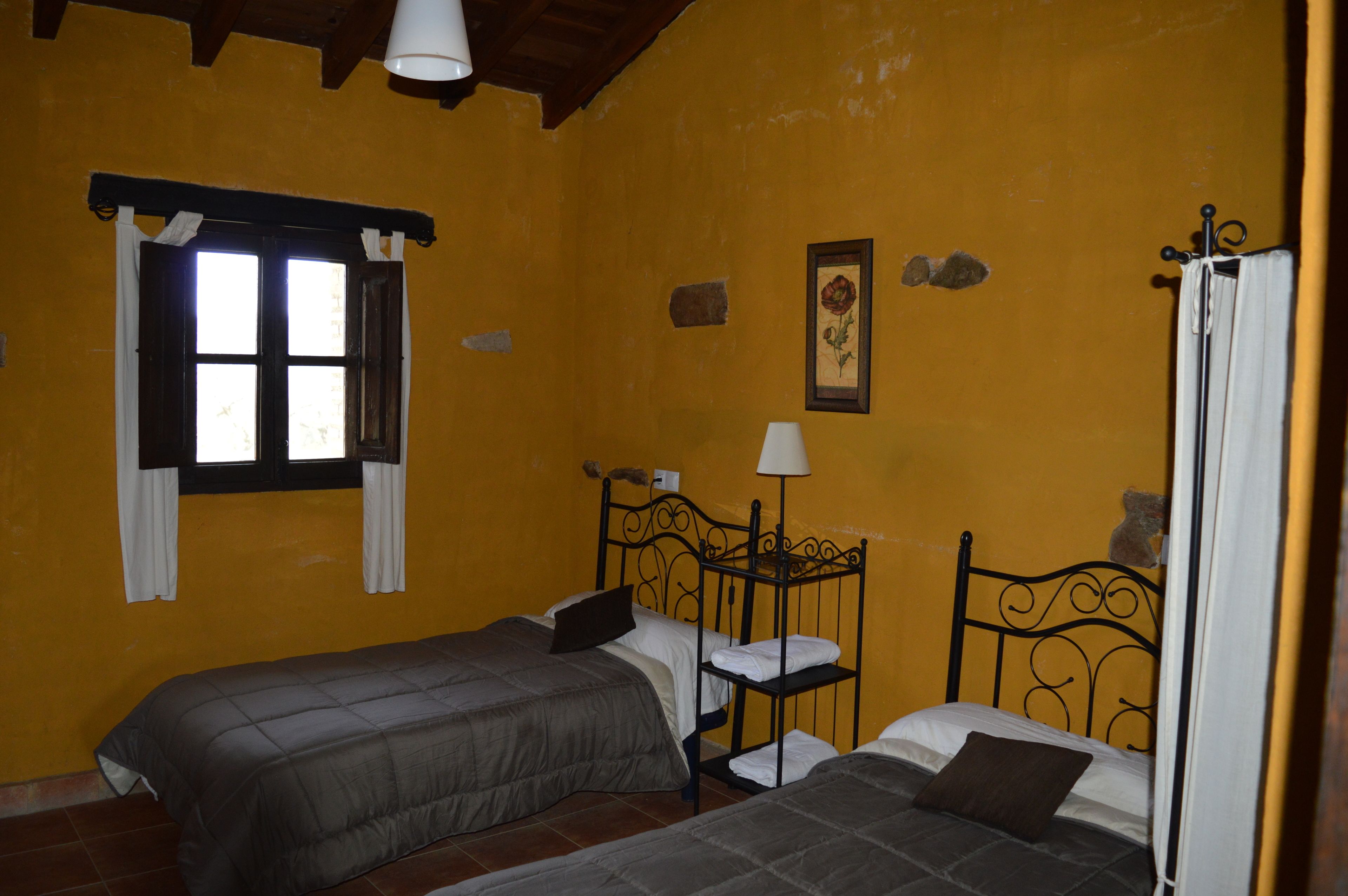 A room with two beds, a window with curtains, a wall-mounted lamp, and a framed picture.