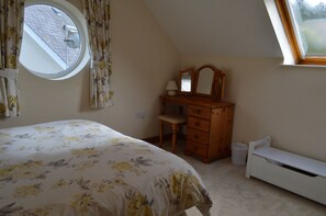Une chambre à coucher comprenant un lit, une commode, une chaise, une petite table avec une lampe, une fenêtre et un radiateur.
