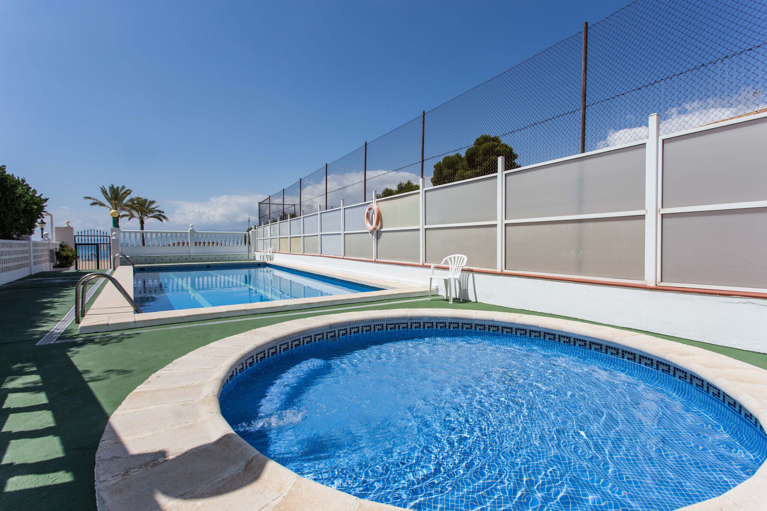 Une piscine dont le bord est recouvert de carreaux, une clôture blanche et une bouée de sauvetage fixée au mur.