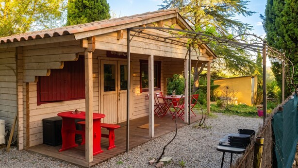 Een houten hut met een veranda, een kleine tafel met stoelen en een barbecue.