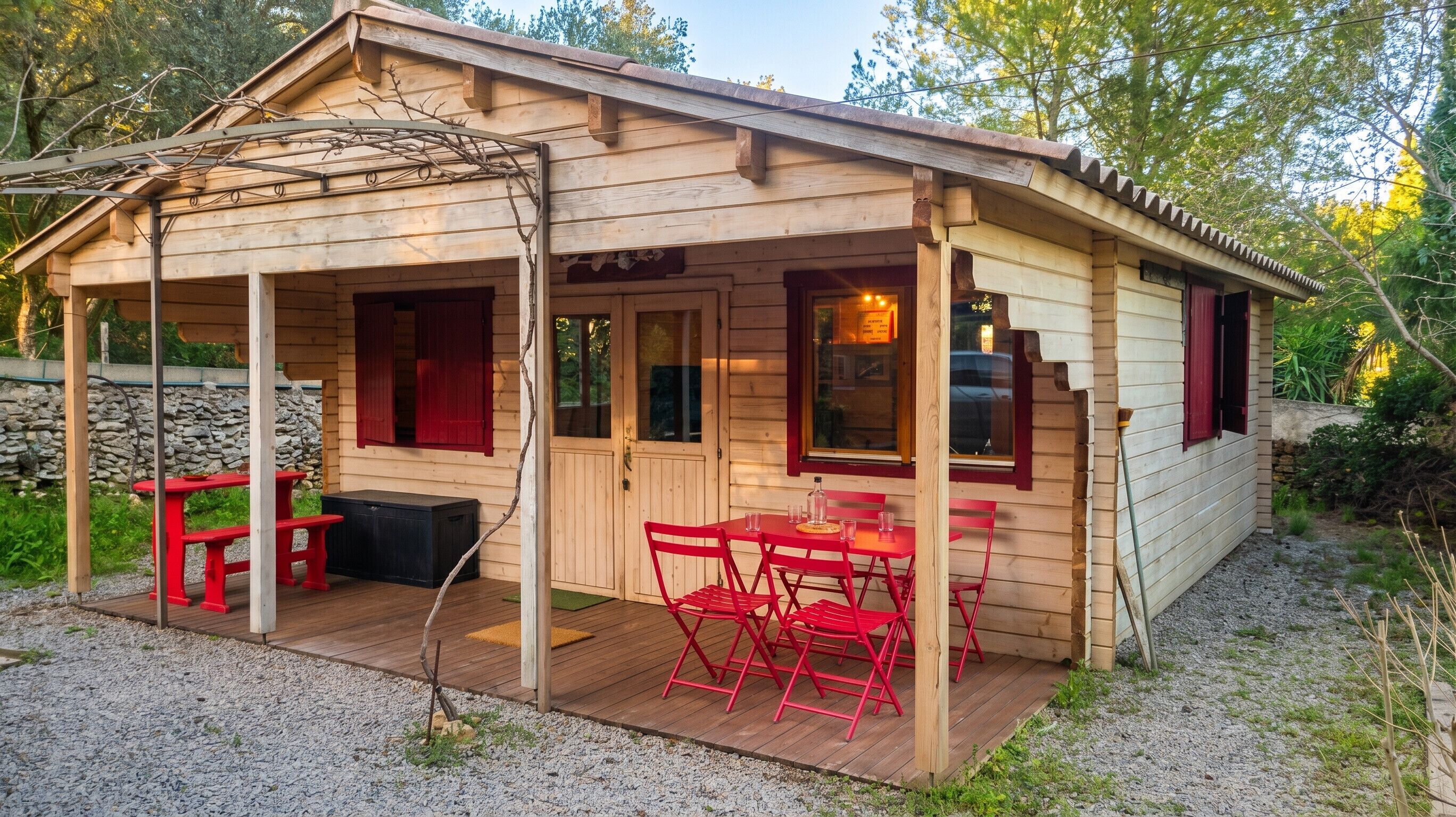 A wooden cabin with a covered patio, a stone wall, and a gravel ground.