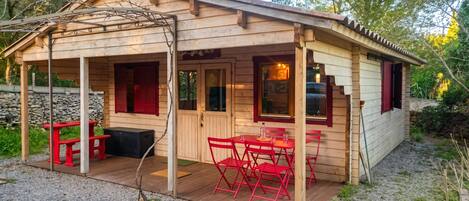 A wooden cabin with a covered patio, a stone wall, and a gravel ground.