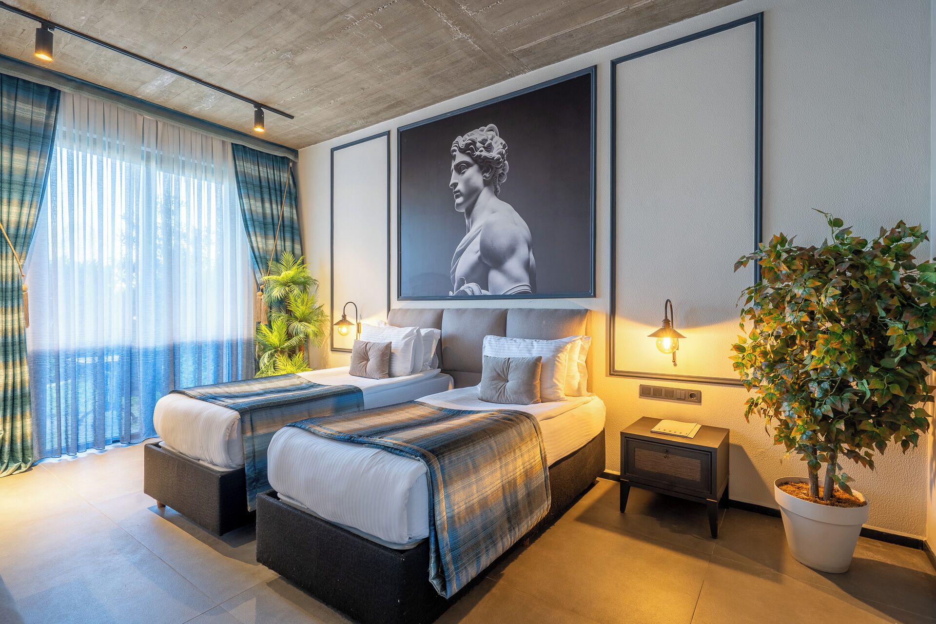 Una habitación de hotel moderna con una cama grande, dos mesitas de noche, una planta en maceta y una obra de arte enmarcada en la pared.