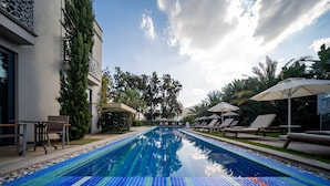 Una piscina con bordes de azulejos, rodeada de sillas de descanso y sombrillas, con un edificio y árboles al fondo.
