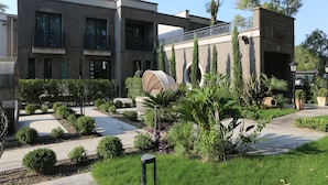 Un edificio moderno con un patio que cuenta con setos podados, un elemento acuático central y un camino que lleva a la entrada.