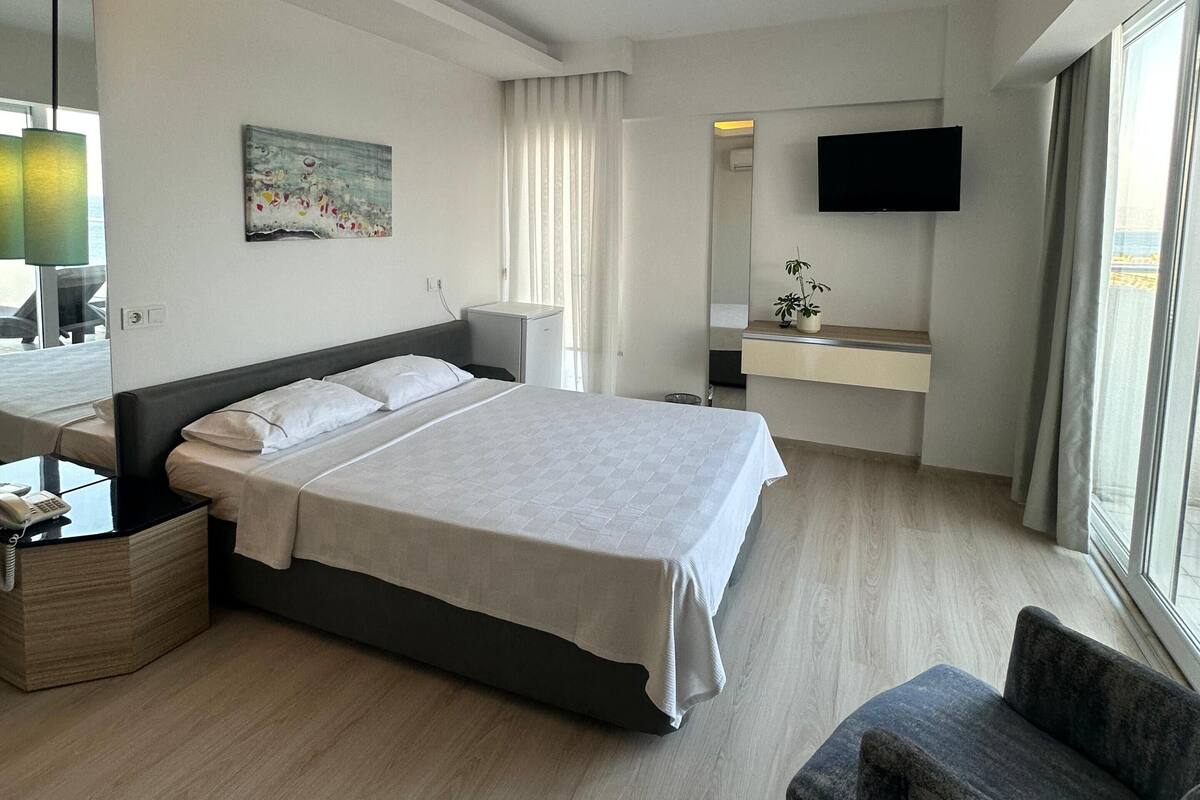 Ein Hotelzimmer mit einem großen Bett, einem Nachttisch mit Telefon, einem Sessel, einem Schreibtisch mit Spiegel, einem an der Wand montierten Fernseher und einem Fenster mit Vorhängen.
