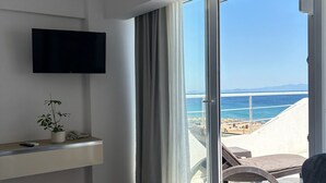 A hotel room with a bed, a TV, a plant, a desk, and a chair. The room has a large window with a view of the beach and ocean. - Amphora Hotel (Ayvalik)