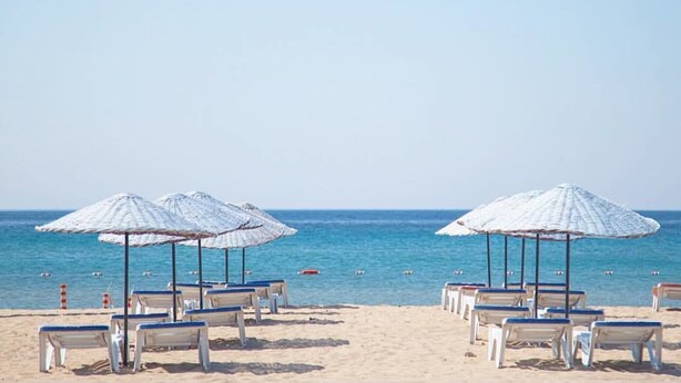 A beach with rows of white lounge chairs and umbrellas, a clear blue sky, and calm sea. - Amphora Hotel (Ayvalik)