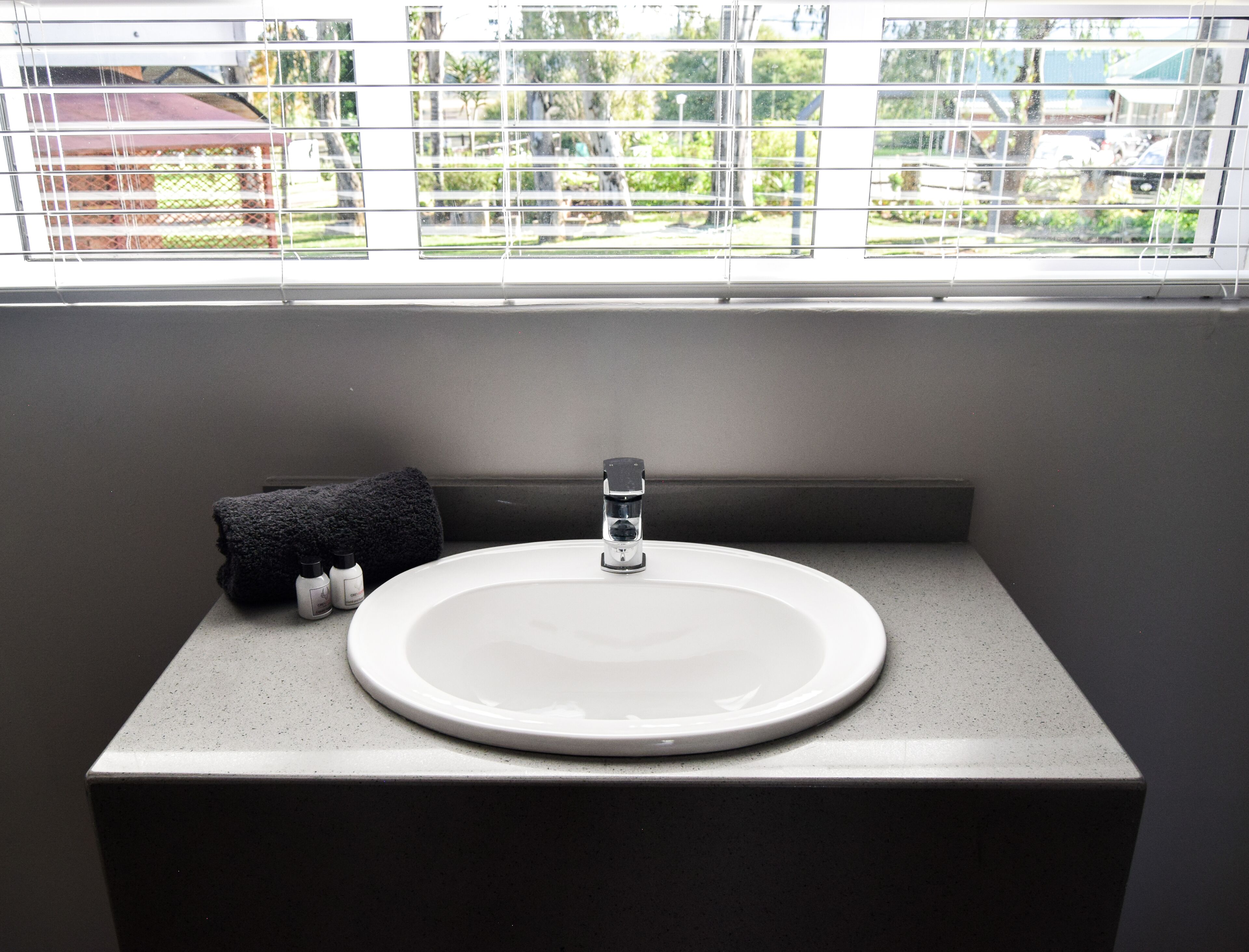Un baño con un lavamanos blanco, una toalla negra y una ventana con persianas.
