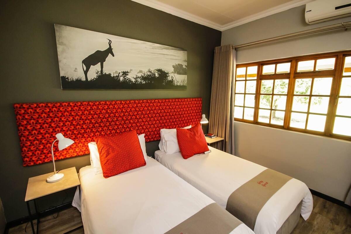 A hotel room with two beds, a red patterned headboard, a painting of an animal, a window with curtains, and an air conditioning unit.