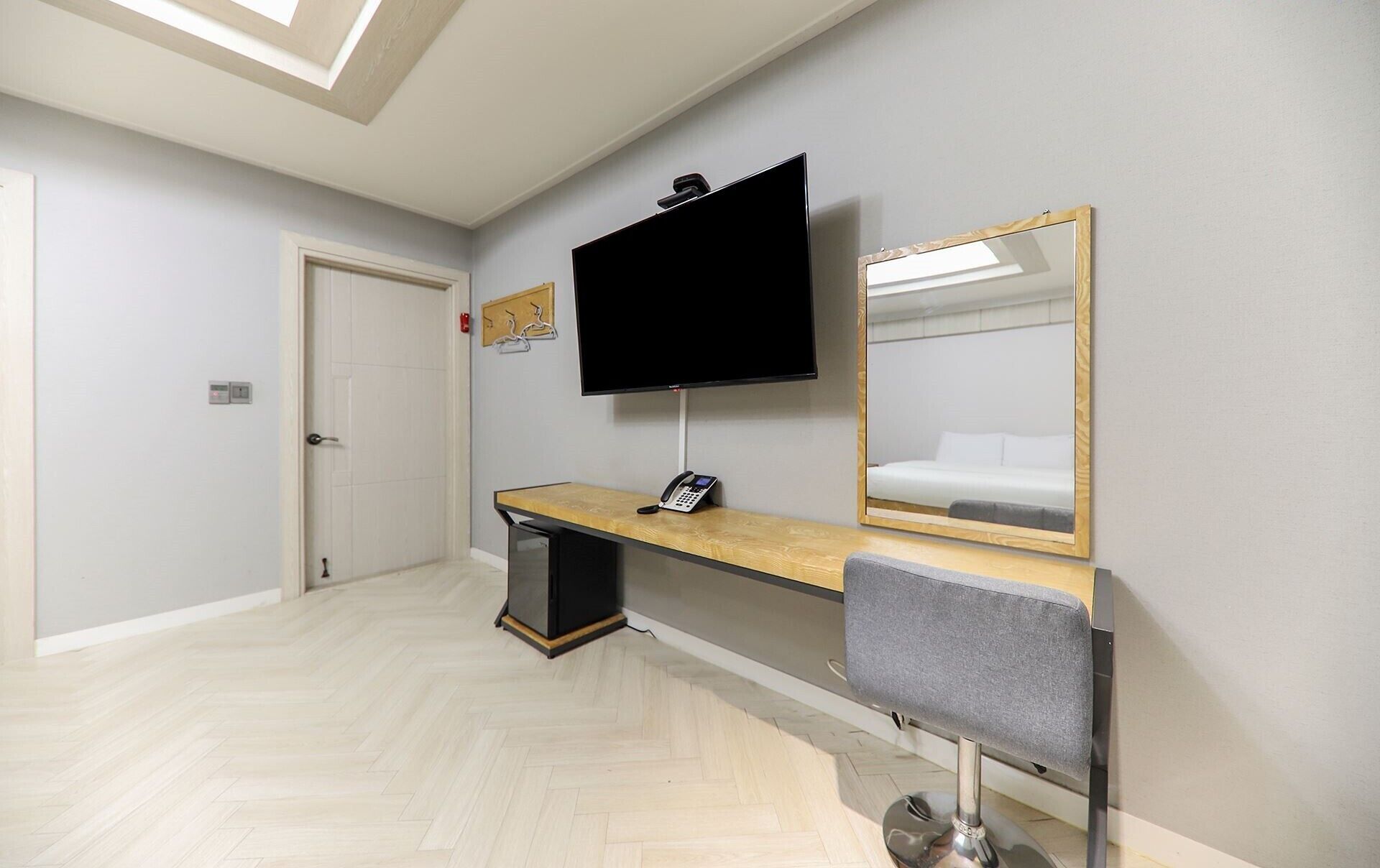 Habitación de hotel con televisor de pantalla plana montado en la pared, un escritorio de madera con teléfono, un espejo y una silla gris.