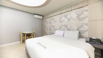 A hotel room with a bed, a small table with a chair, a wall-mounted air conditioner, and a TV.