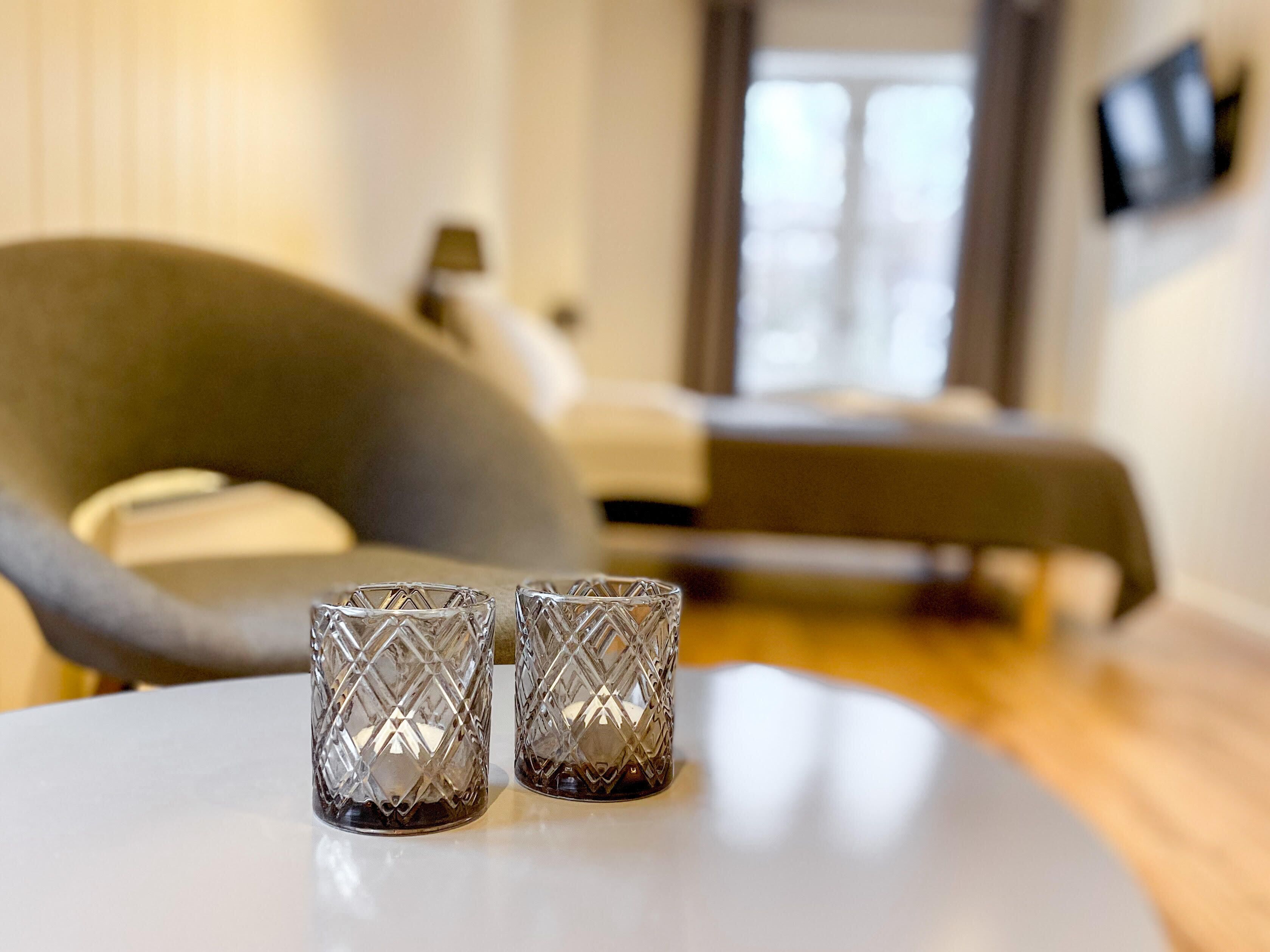 Quarto de hotel moderno com uma cama, uma poltrona cinza e dois vasos de vidro decorativos sobre uma mesa redonda.