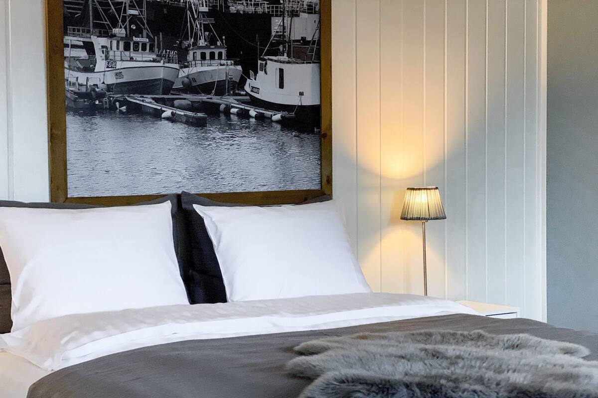 Een bed met wit en grijs beddengoed, een bont plaid en een lijst met een foto van boten aan de muur.