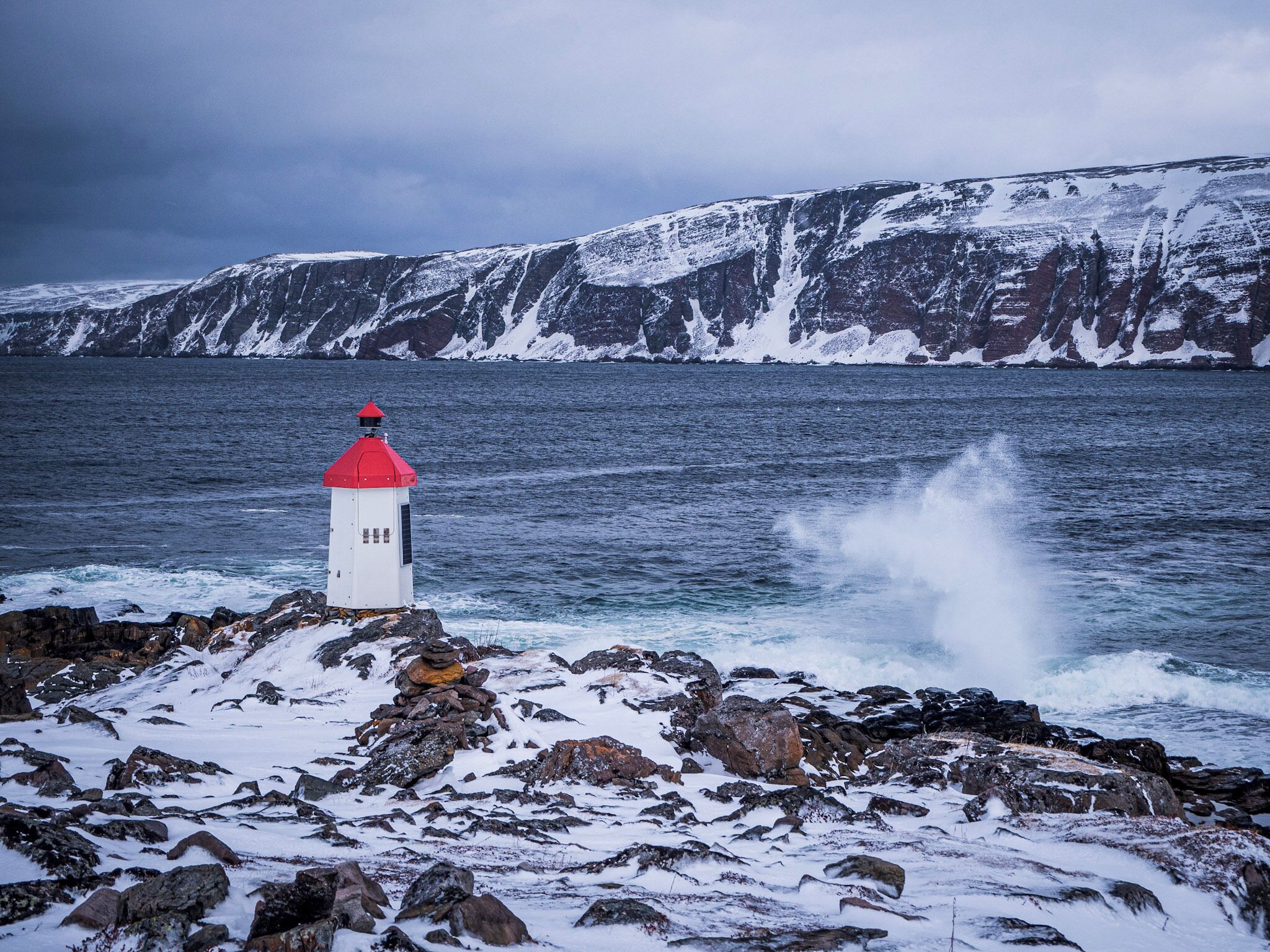 A lighthouse on a rocky shore with snow-covered cliffs in the background.