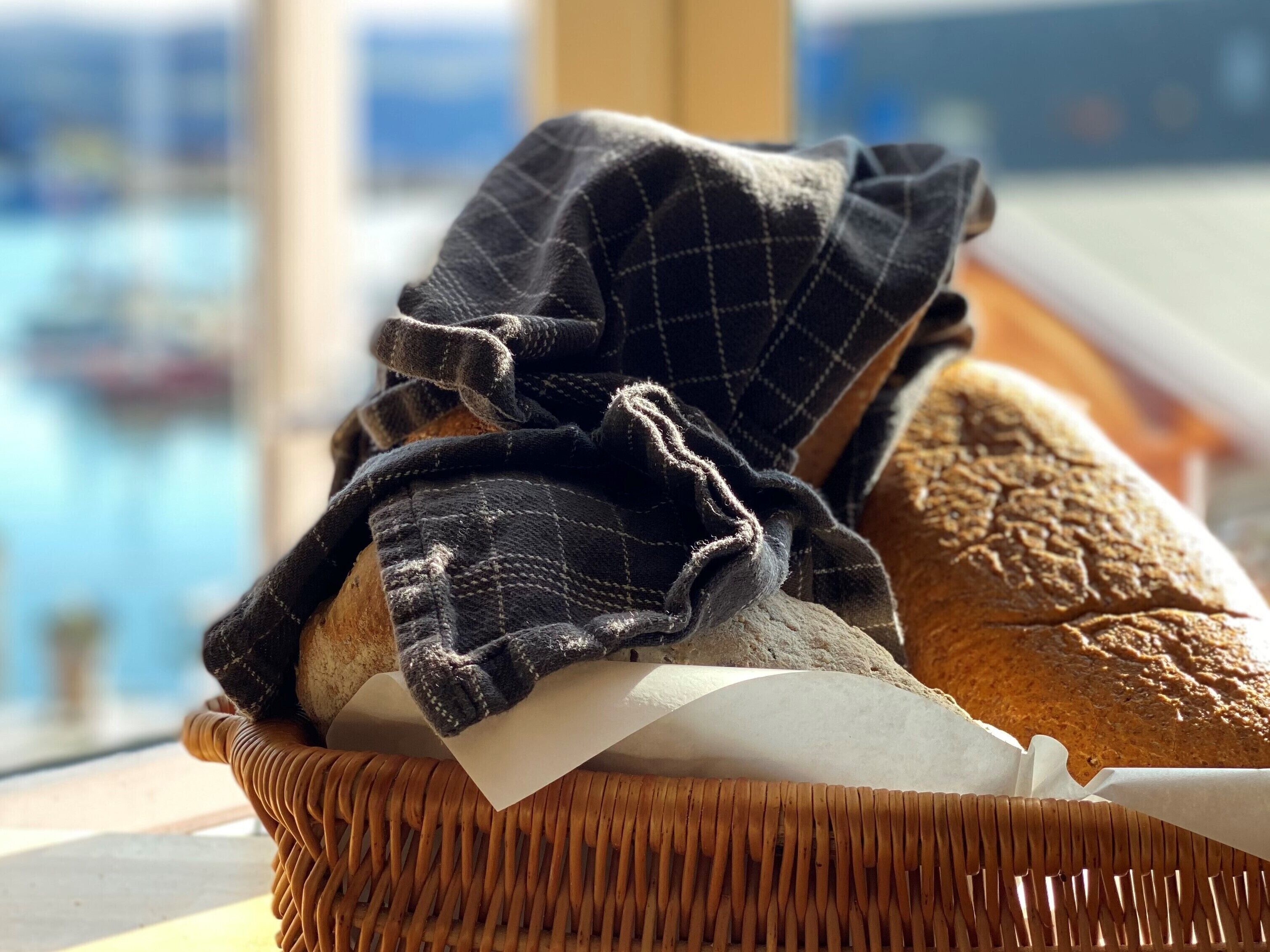 Un panier en osier contenant du pain et une étoffe, posé sur une table près d’une fenêtre donnant sur un plan d’eau.