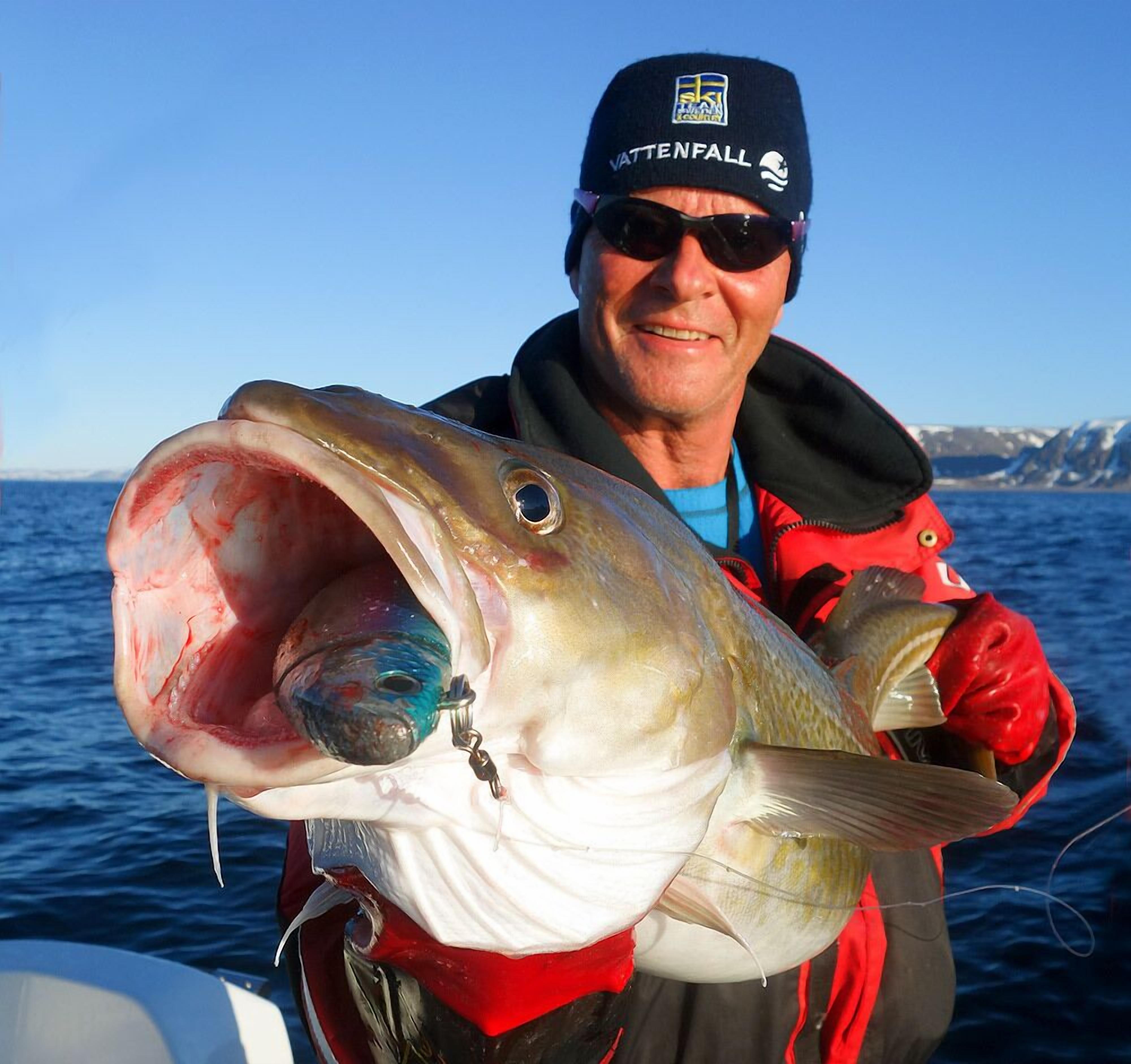 A person holding a large fish with a hook in its mouth, wearing a jacket with a logo and a beanie.