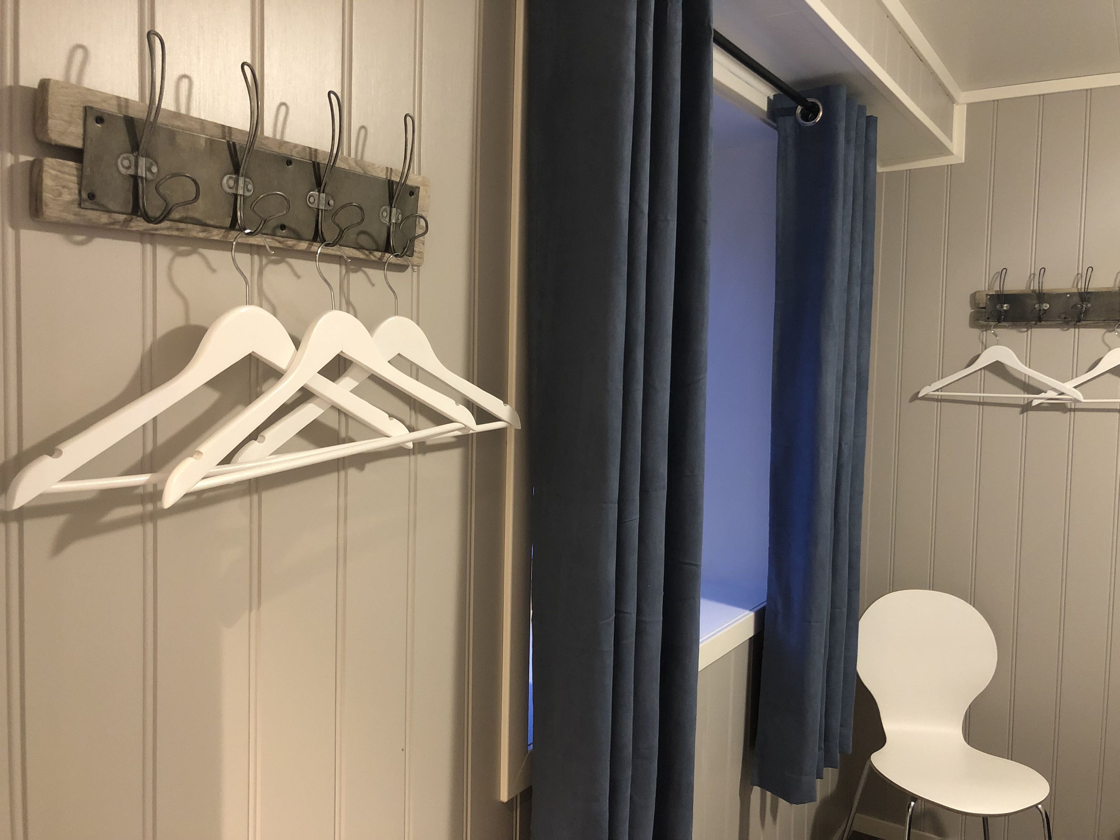 Ein Zimmer mit einer Holzwand, einem Fenster mit blauen Vorhängen, einem weißen Stuhl und einem Kleiderständer mit weißen Kleiderbügeln.