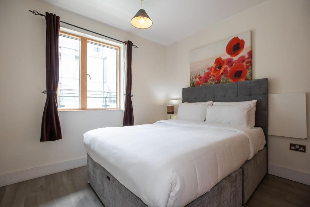 A bedroom with a bed, a window with curtains, a painting, and a lamp.