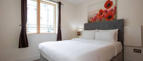 A bedroom with a bed, a window with curtains, a painting, and a lamp.