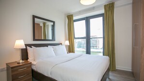 Ein Hotelzimmer mit einem großen Bett, einem Nachttisch mit Lampe, einem Spiegel und Blick auf eine Stadtlandschaft durch das Fenster.