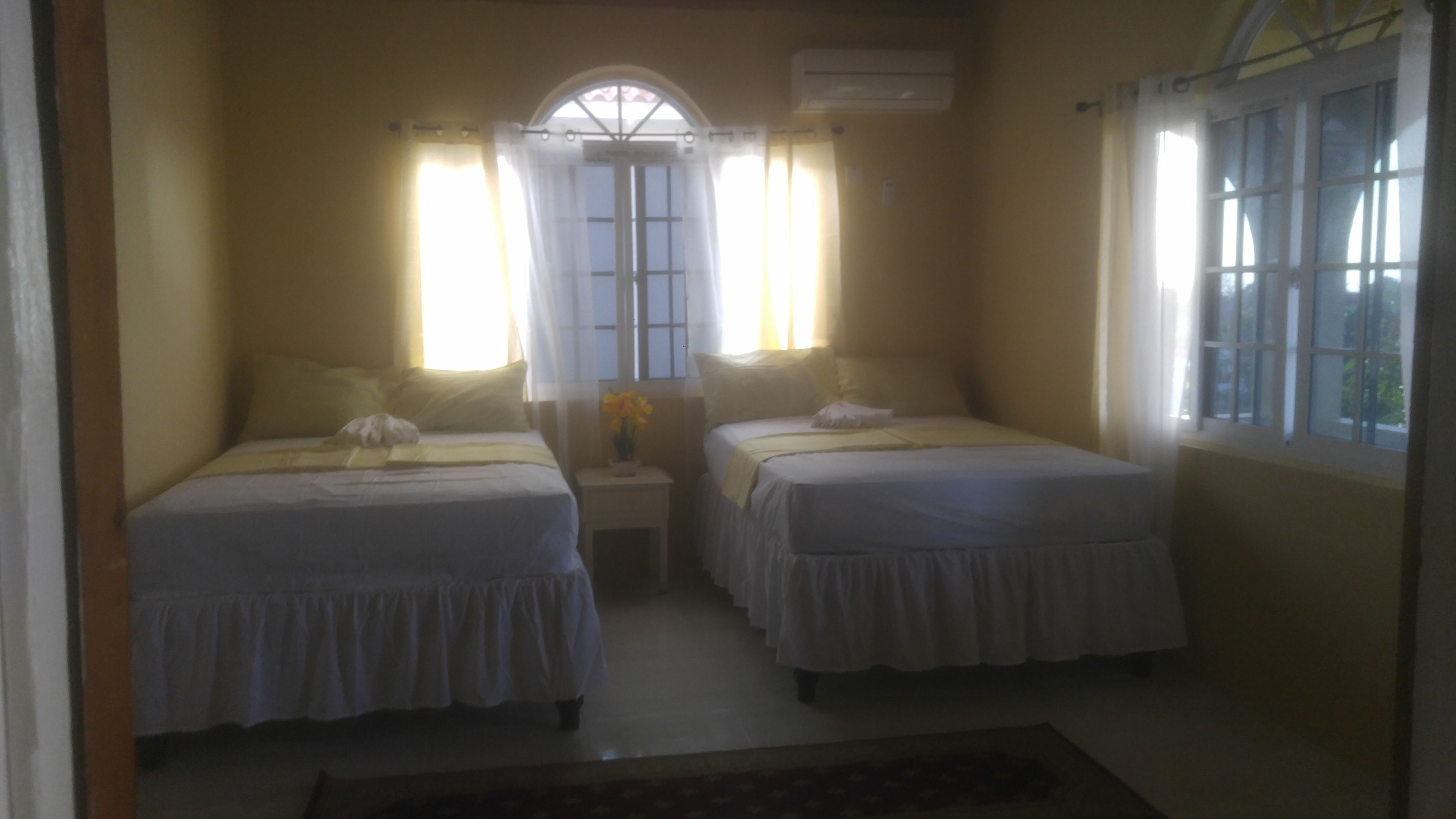 A room with two beds, a window with curtains, an air conditioning unit, and a small table with a vase of flowers.