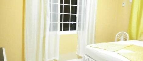A bedroom with a bed, white curtains, a window, and an air conditioning unit.