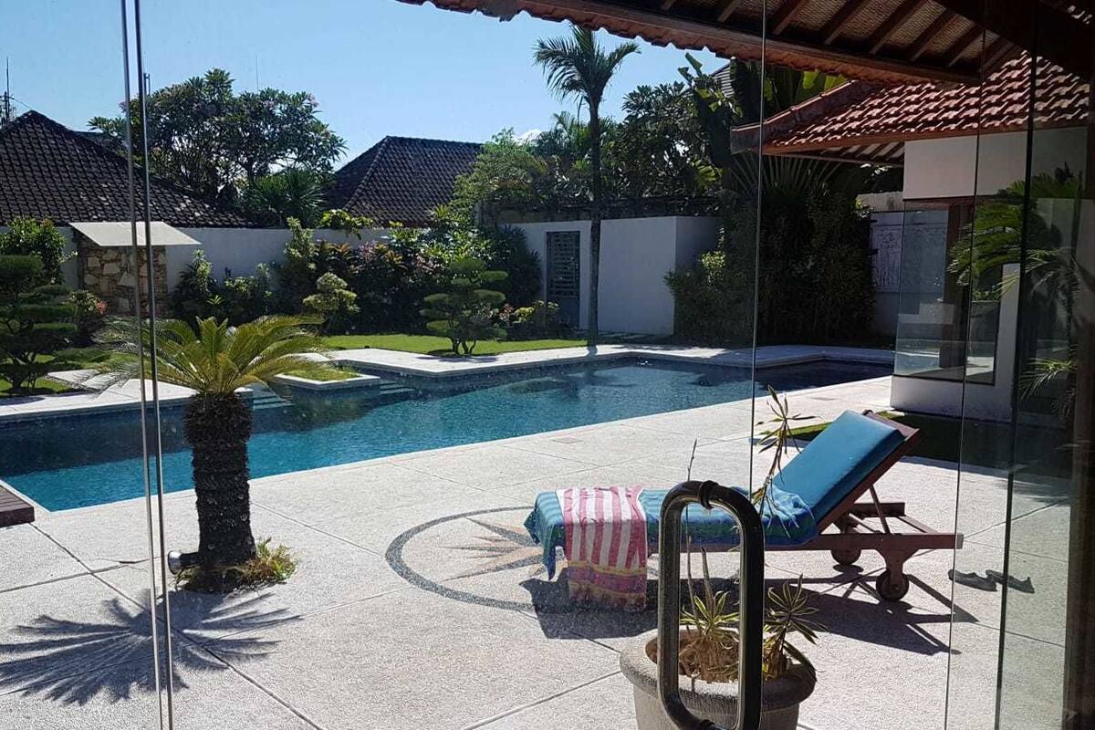 A swimming pool with a lounge chair and a small table, surrounded by a garden and a building with a tiled roof.