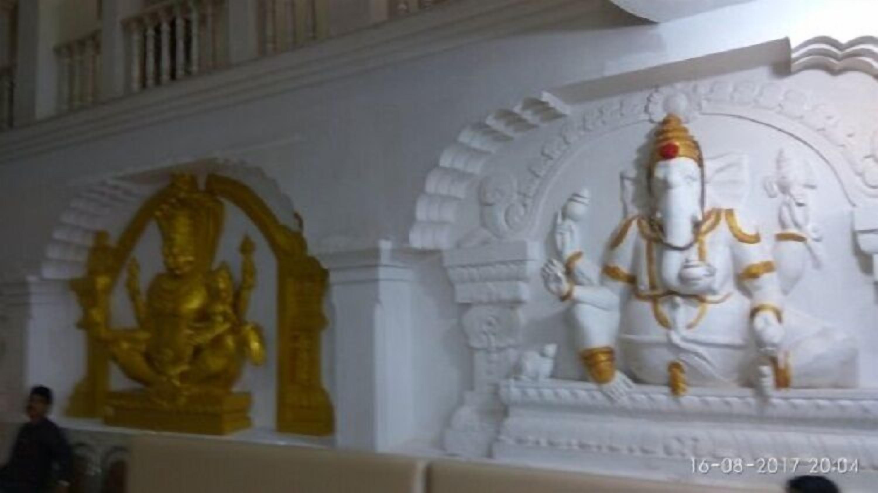 A statue of a deity with a golden halo and a white figure with golden accents, set against a white wall with decorative arches.