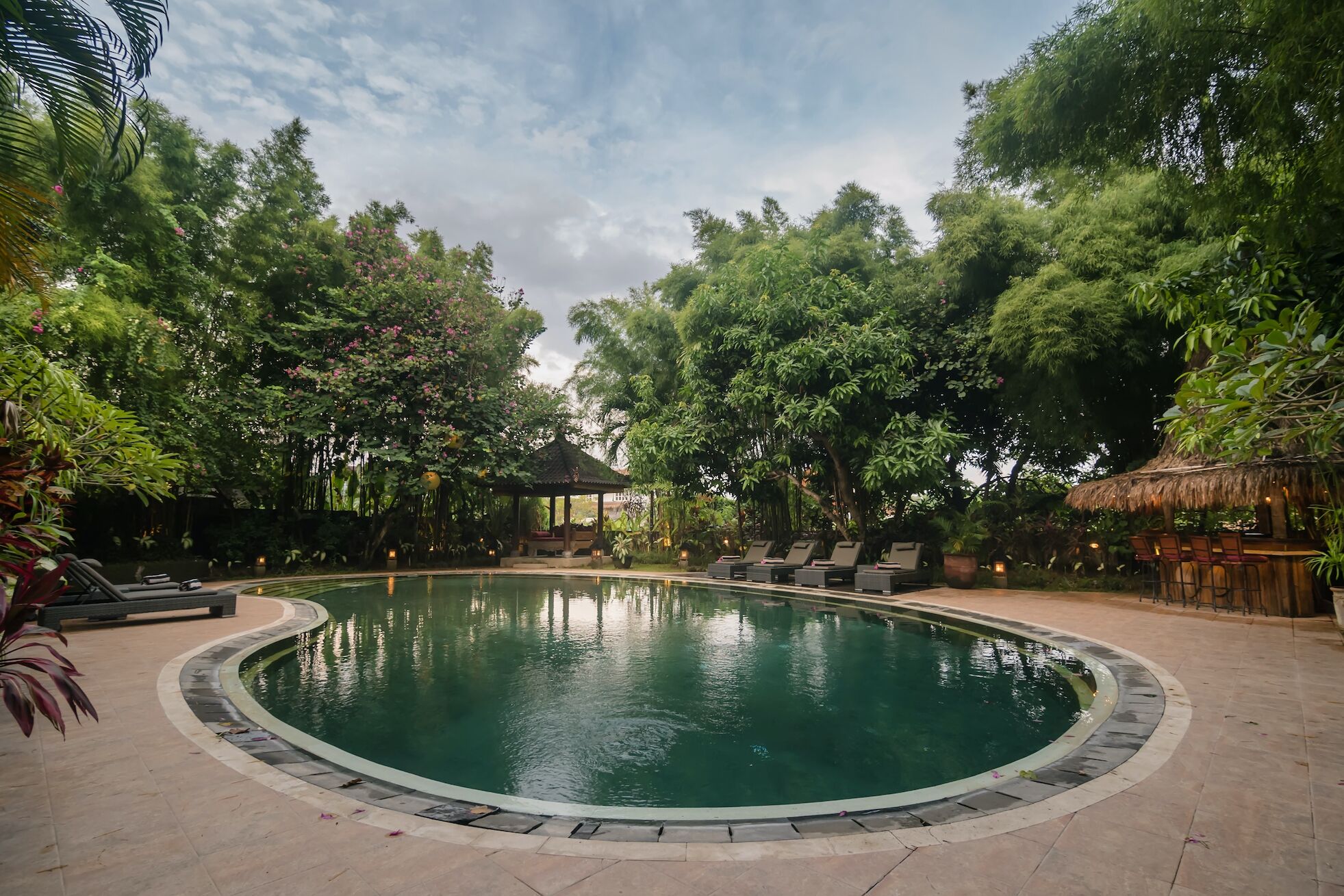 Una piscina circular rodeada de sillones de descanso y un pabellón, con abundante vegetación y árboles al fondo.