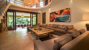 A spacious living area with a large sectional sofa, a wooden coffee table, and a dining area with chairs. A staircase with a railing leads to an upper level, and a large abstract painting is visible on the wall. - Large Groups,9 BDR,Close To Beach,Great Inclusions (Canggu)