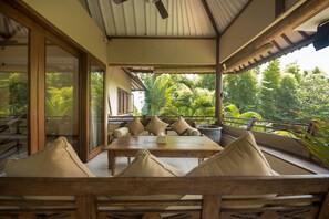 A covered outdoor seating area with a wooden table and chairs, surrounded by lush greenery and a view of the garden.