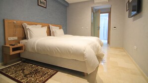 A bedroom with a large bed, a nightstand, a TV mounted on the wall, and a door leading to another room. - Hotel Kenzo (Safi)