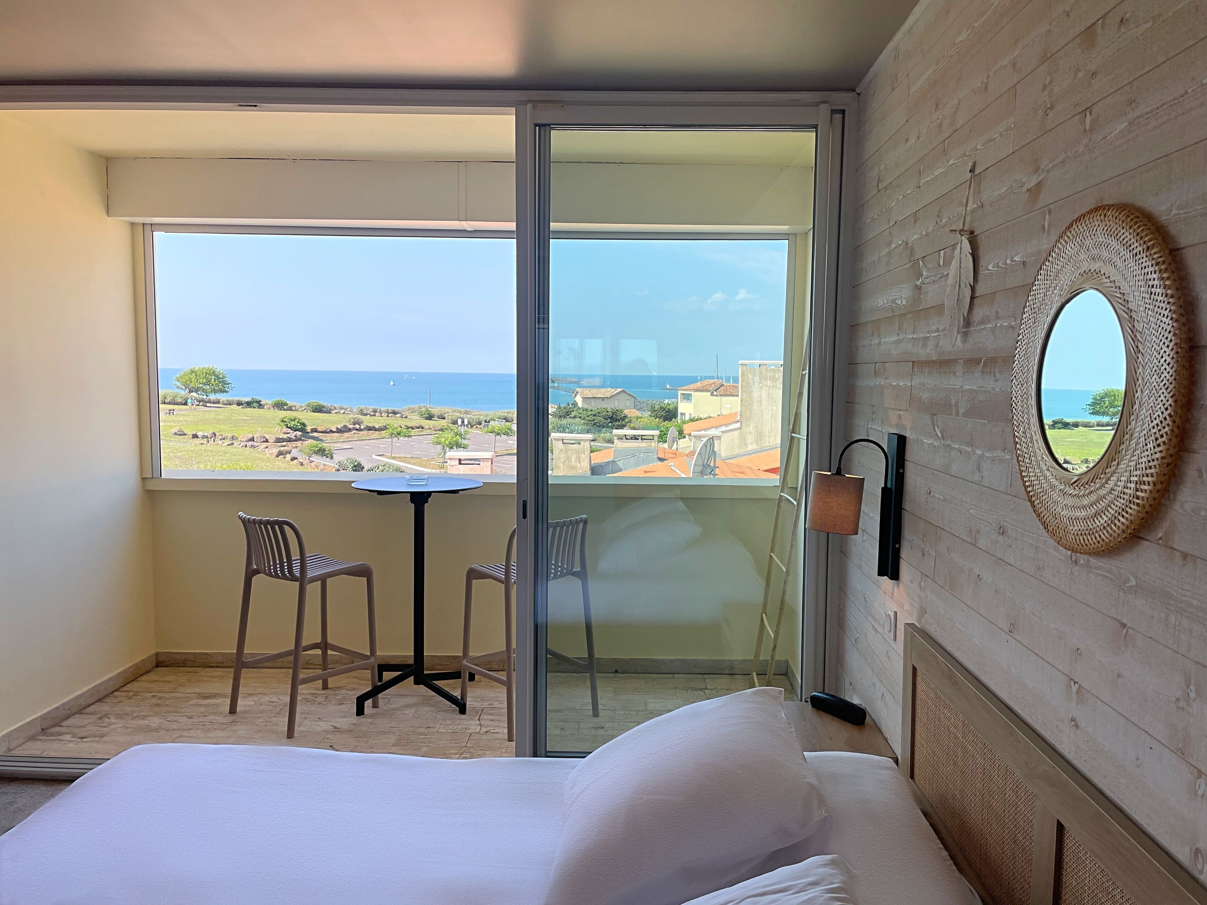 A balcony with a table and chairs, a bed with white linens, and a view of the sea and buildings.