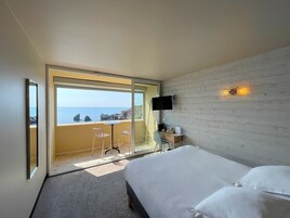 A bedroom with a bed, a desk, a chair, a TV, and a balcony with a view of the sea.