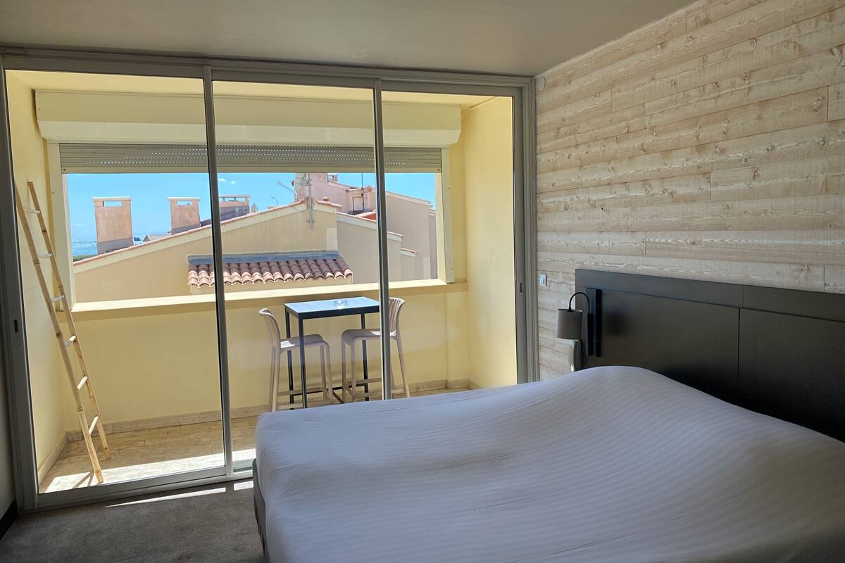 A bedroom with a bed, a small table with chairs, a window with a view of buildings, and a wooden wall.