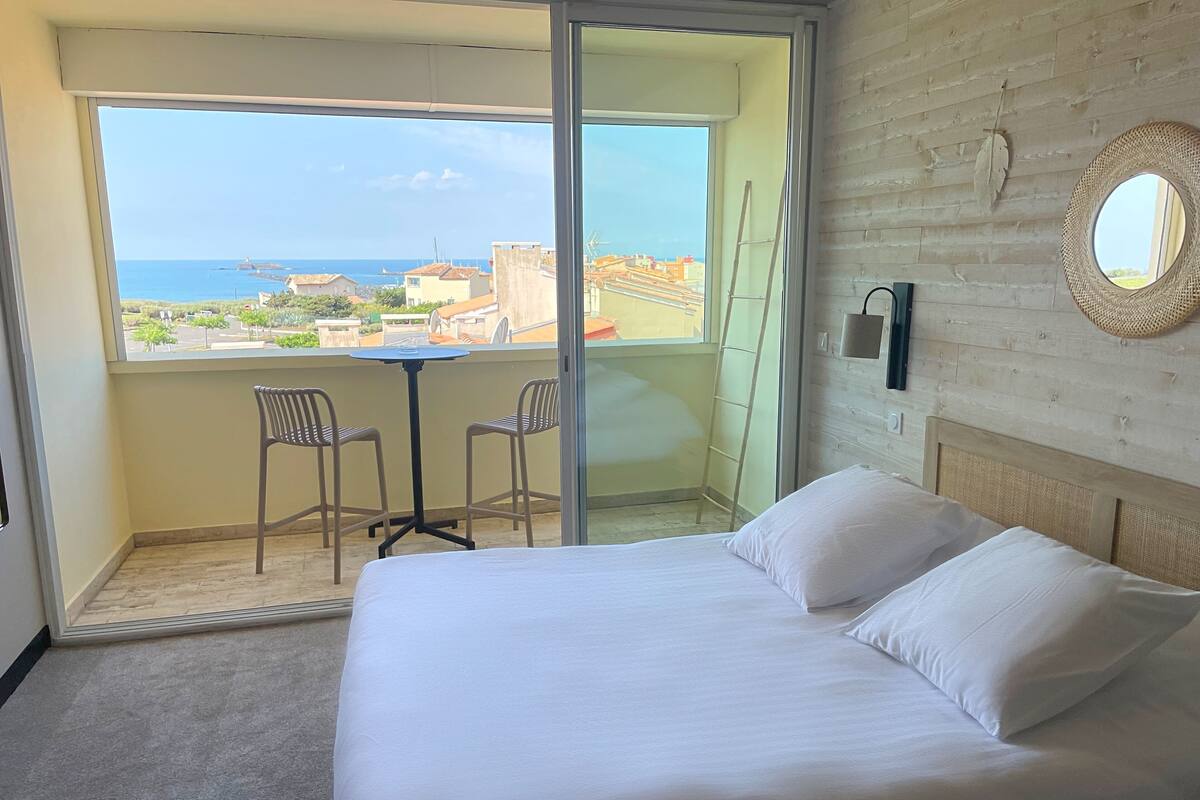 A hotel room with a bed, a small table, two chairs, a view of the sea, and a balcony.