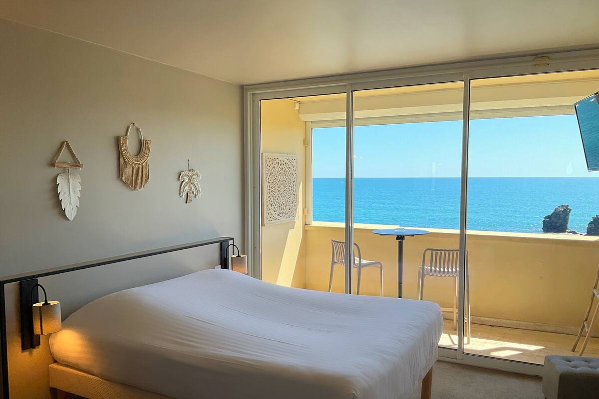 A bedroom with a bed, a small table, two chairs, and a view of the sea through a sliding glass door.