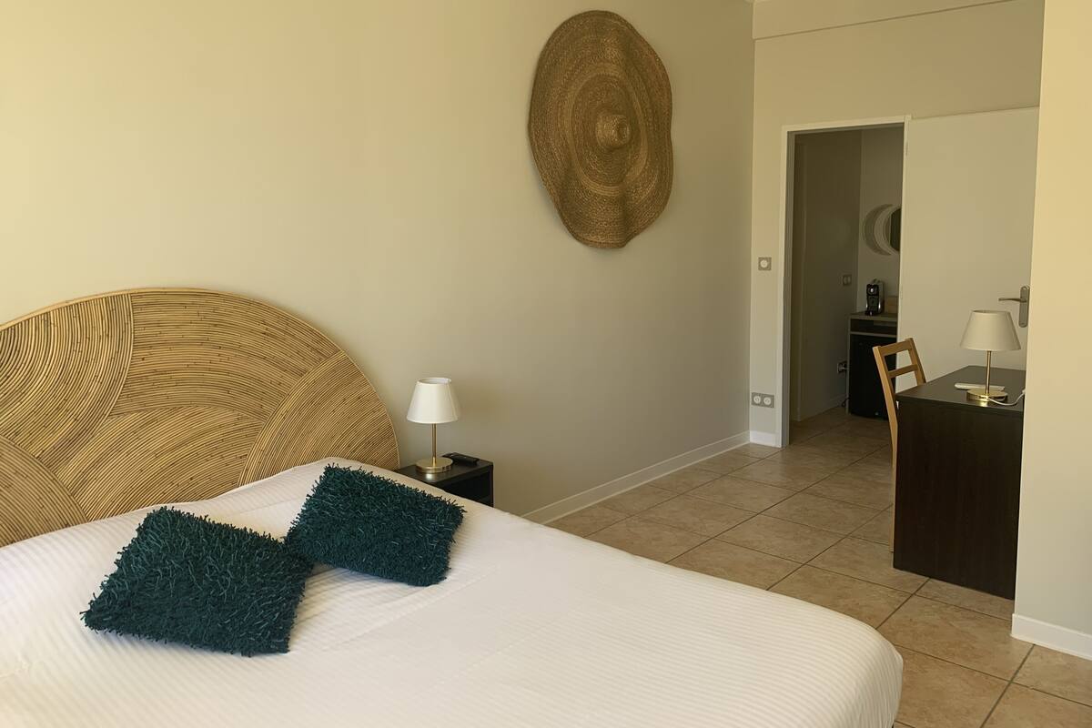 A neatly made bed with a white comforter and two dark green pillows, a round woven wall art piece, a small bedside table with a lamp, and a dark wooden desk with a lamp in a room with beige walls and tiled flooring.