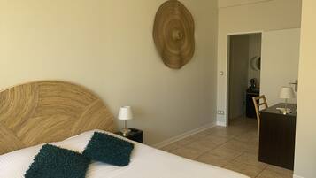 A neatly made bed with a white comforter and two dark green pillows, a round woven wall art piece, a small bedside table with a lamp, and a dark wooden desk with a lamp in a room with beige walls and tiled flooring.