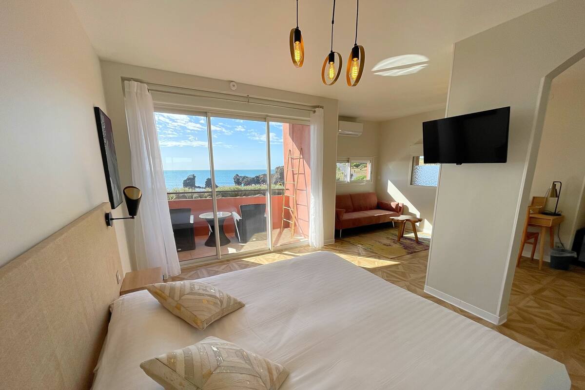 A hotel room with a large bed, a sofa, a TV, and a balcony with a view of the sea.