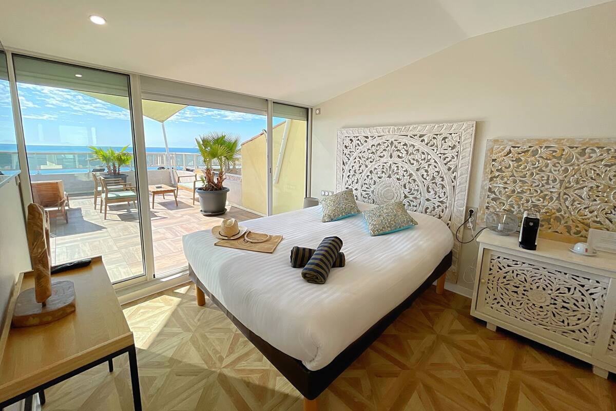 A bedroom with a large bed, a view of the sea, a balcony with chairs and a table, and a decorative headboard.
