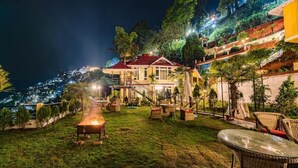 Área externa bem iluminada com mesa e cadeiras, churrasqueira e vista para uma encosta com edifícios.